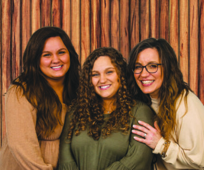 three women with long hair posing together in front of wooden backdrop