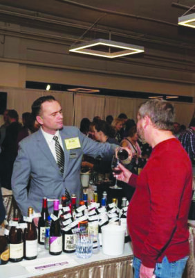 man wearing suit serving wine samples behind table at indoor wine event