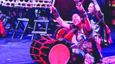 Asian women sitting behind large drums, arms raised to play, on stage during performance