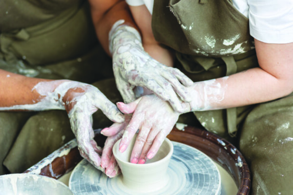 two sets of hands, covered in clay, one guiding the other in making a pot on a pottery wheel