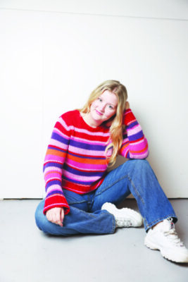 A young woman poses seated on the ground in a striped red and pink sweater.