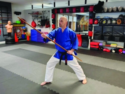 Lenny Demers poses in a long fighting stance with a wooden staff in his studio.