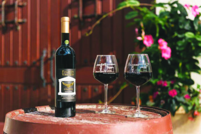 bottle of wine sitting on barrel beside quarter full stemmed wine glasses, outside with flowers and wooden door in background