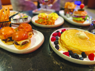 A table of delicious food, including mini burgers, a large pretzel, and pancakes.