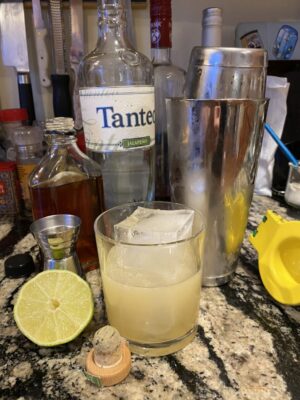 ball glass with ball glass containing light colored cocktail over ice, surrounded by ingredients and cocktail making equipment.