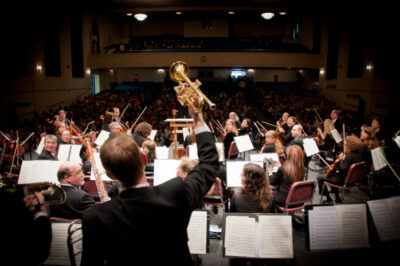 members of an orchestra seen from the back, trumpet player holding up his trumpet in one hand, dark stage