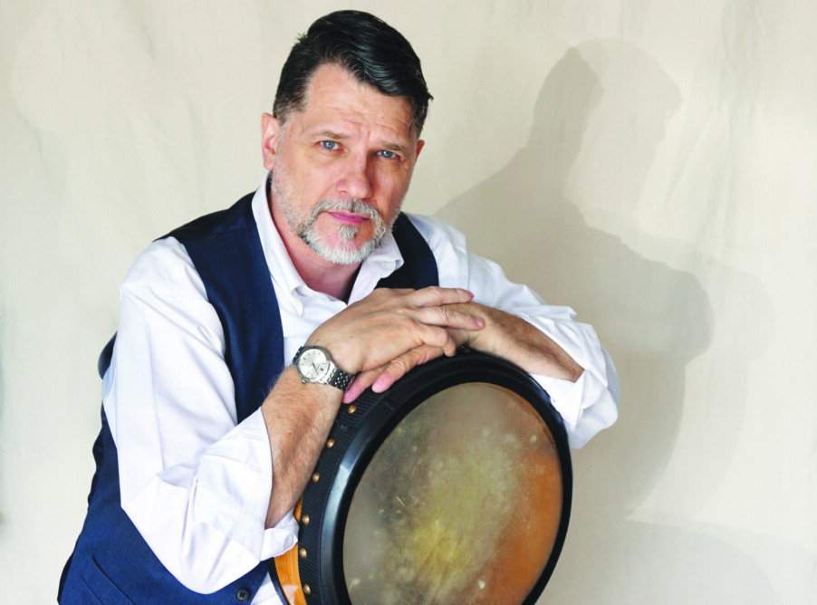 middle aged white man with dark hair and gray beard, wearing white shirt with dark vest, leaning on hand drum, portrait style