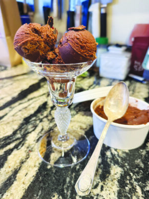 two scoops of chocolate sorbet in stemmed glass, sitting on counter beside cardboard cup of ice cream with spoon