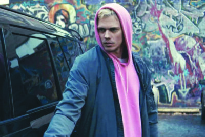 Bill Skarsgard wearing a pink hoodie and dark jacket, standing next to car in graffiti covered alley, in screenshot from movie Locked