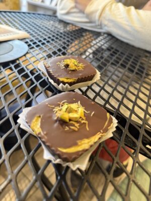 large squares of filled chocolate topped with pistachios, sitting in paper wrappers on metal table