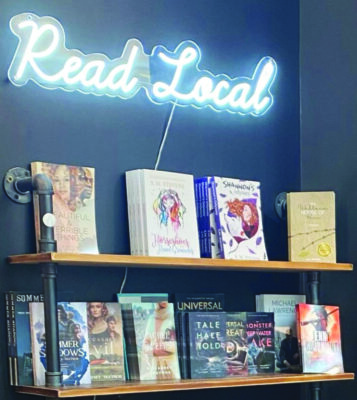 shelves along dark painted wall holding faced out books, neon light above reading "Read Local"