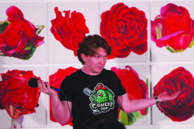 man standing in front of wall with large photos of red roses, holding microphone and making shrugging gesture