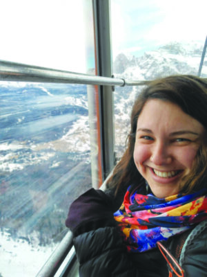 head shot of woman standing beside window that looks out on mountain landscape with snow on the ground, woman dressed in cold weather clothes, smiling