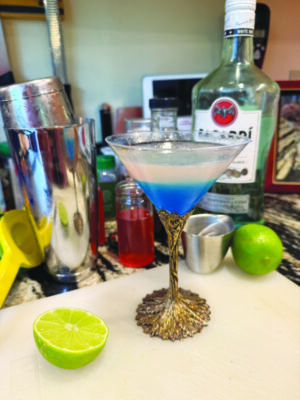 fancy martini glass filled with cocktail that is blue at the bottom and white at the top, sitting on counter with limes and cocktail shaker