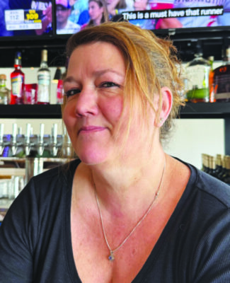 white woman bartender in front of bar shelves, closed mouth smile, looking slightly mischievious