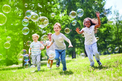 A group of kids run and smile in a field as they chase bubbles