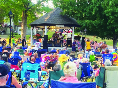 crowd of people sitting in folding chairs in park around bandstand where band is playing