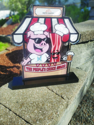 trophy with flat cut out image of pig wearing chef's heat and sunglasses hanging out with piece of bacon, also wearing sunglasses, both behind counter with stripped awning