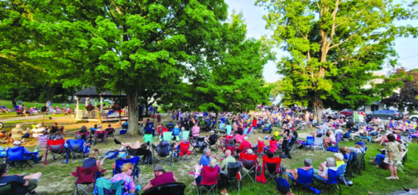wide view of crowd of people sitting in folding chairs in loose groups around a bandstand. Evening in summer