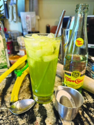 tall glass with green colored cocktail, filled with ice, sitting on counter amongst cocktail making equipment