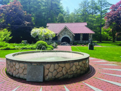 round stone pool with small fountain spigots on brick paved area of grassy park, brick path leading to stone building behind fountain, trees and garden beds