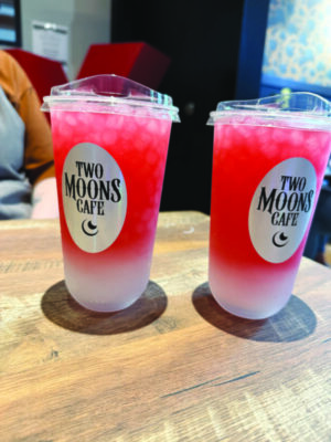 two clear plastic take-out cups with lids, sitting on wooden table. Filled with lemonade that gradiates from dark pink to white at the bottom.