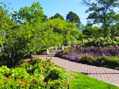brick walkway through park with cultivated garden beds filled with colorful flowers, small white fence containing large garden area