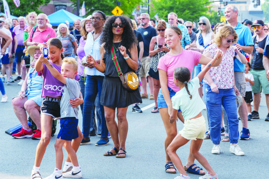 crowd of people standing on pavement, kids at the front dancing, dusk on summer day