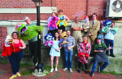 group of performers standing in front of a brick wall and lamp post, most of them holding puppet characters