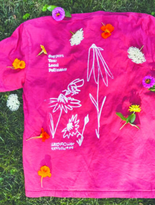 dark pink shirt laid out on grass, seen from above, with words "Support Your Local Pollinators" and drawings of flowers printed on it