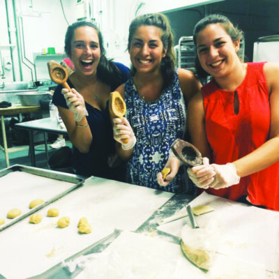 three young women standing behind table with cookie dough, they are smiling and holding up wooden cookie molds