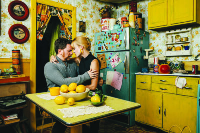 a man and a woman wrapped together in an intimate embrace at a small table in a kitchen with vintage decor, bowl of lemons on the table