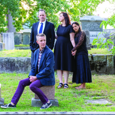 actors posing in a graveyard, one man sitting on a headstone in foreground, in background a man and two women holding hands to comfort each other. Melancholy feel