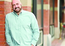 bald man with beard wearing button up shirt, leaning against brick building on downtown sidewalk