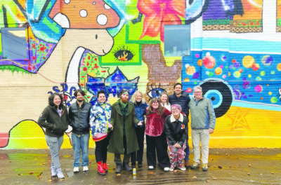 group of people standing on sidewalk in front of large painted mural on brick building, colorful mural of collage of drawings