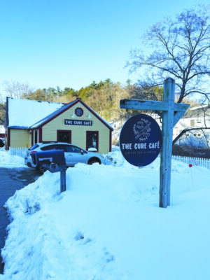sign hanging from wooden post on snow covered lawn, in front of cozy looking building