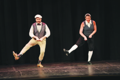 a man and a woman, dressed in vests and newsboy caps dancing in sync on dark stage