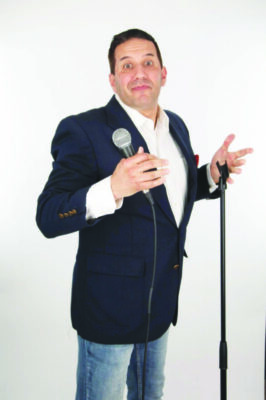 white man dressed in jeans, a white button up shirt and blazer, standing in front of microphone stand holding microphone and making shrugging motion