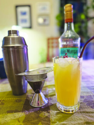 Tall ball glass filled ice and a cocktail, sitting beside cocktail shaker and measuring cup on table