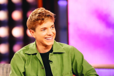 young looking androgynous white person smiling and looking to the side, sitting in talk show studio