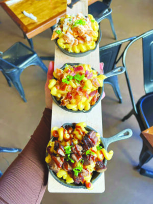 wooden food board holding three small cast iron skillets with macaroni and cheese with different toppings, lined up