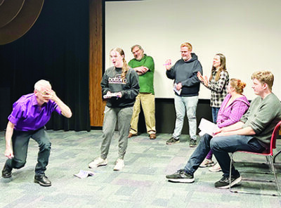 rehearsal of play showing group of people standing in circle, one young woman in the center while a man hunches forward dramatically