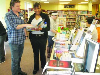 people standing beside tables full of books, looking at one open book during event