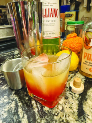 short ball glass filled with red and yellow cocktail and ice, sitting on counter with cocktail shaker and ingredients