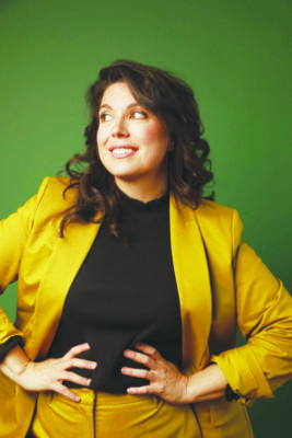 white woman wearing mustard yellow blazer, hands on hips, grinning and looking to the side