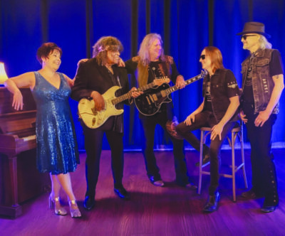 five band members standing in dark room, two holding guitars, woman leaning against piano