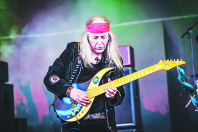 Long haired elder man wearing headband and black coat, standing on stage while playing guitar