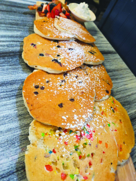 four kinds of pancakes on a wooden board with small pile of fruit and bacon and bowl of whipped cream
