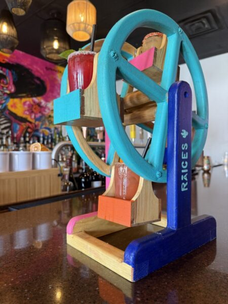 short glasses of different margaritas with salt rims sitting in caddies on a small wooden ferris wheel made for a fun display