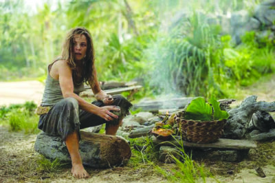 scene from film Send Help with Rachel McAdams sitting on a log beside fire, wearing torn up clothes